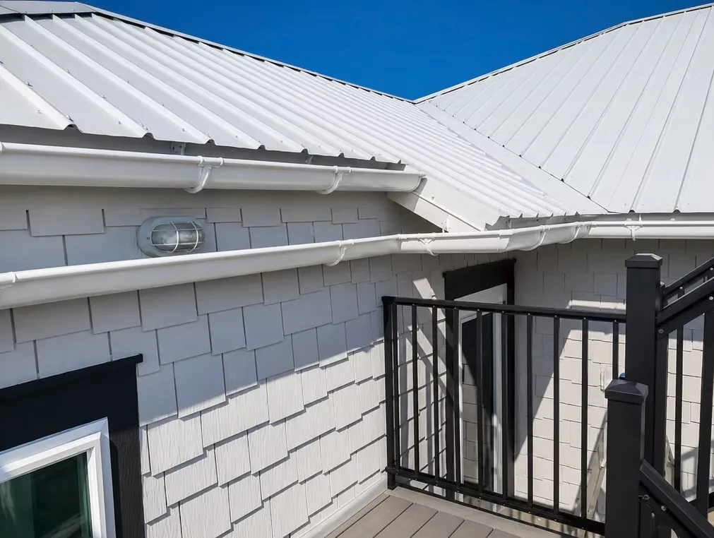 White half-round gutters on round hangers beneath a white standing-seam metal roof, on a grey shake-sided home with a black deck railing