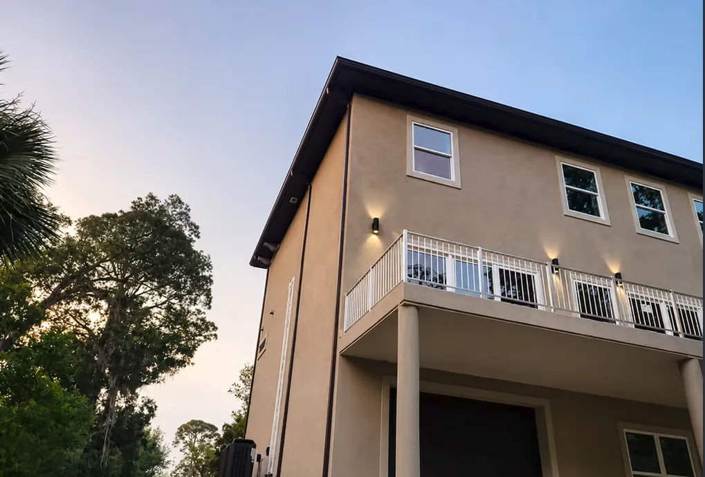 Dark bronze gutter and downspout on a tan stucco two-storey home with a white balcony railing, at dusk