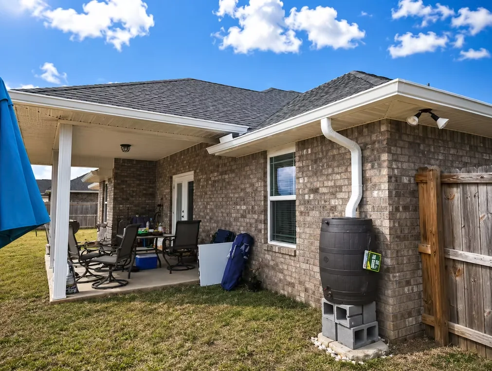 White seamless aluminium gutter and downspout on the back corner of a tan brick single-storey home, feeding a rain barrel beside a covered patio